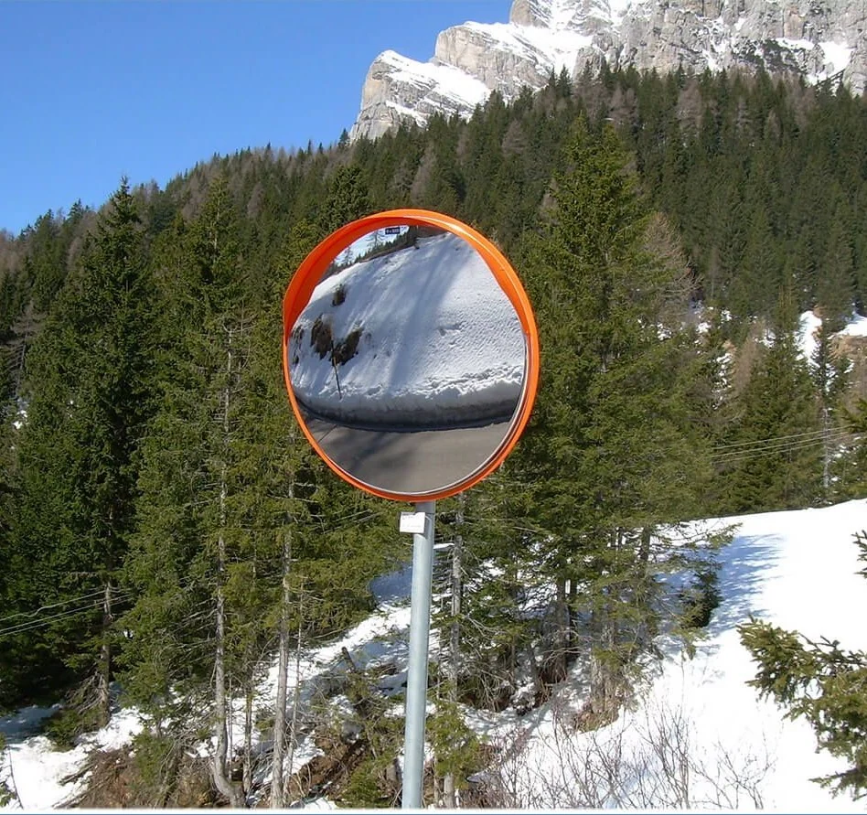 Specchio convesso con bordo arancione su un palo davanti a un bosco e a un pendio innevato.