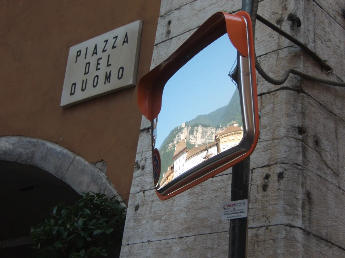 Specchio convesso rettangolare con bordo arancione fissato all’angolo di un edificio in pietra.