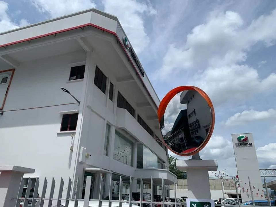 Specchio convesso con bordo arancione montato su un pilastro davanti a un edificio bianco e a una recinzione metallica.