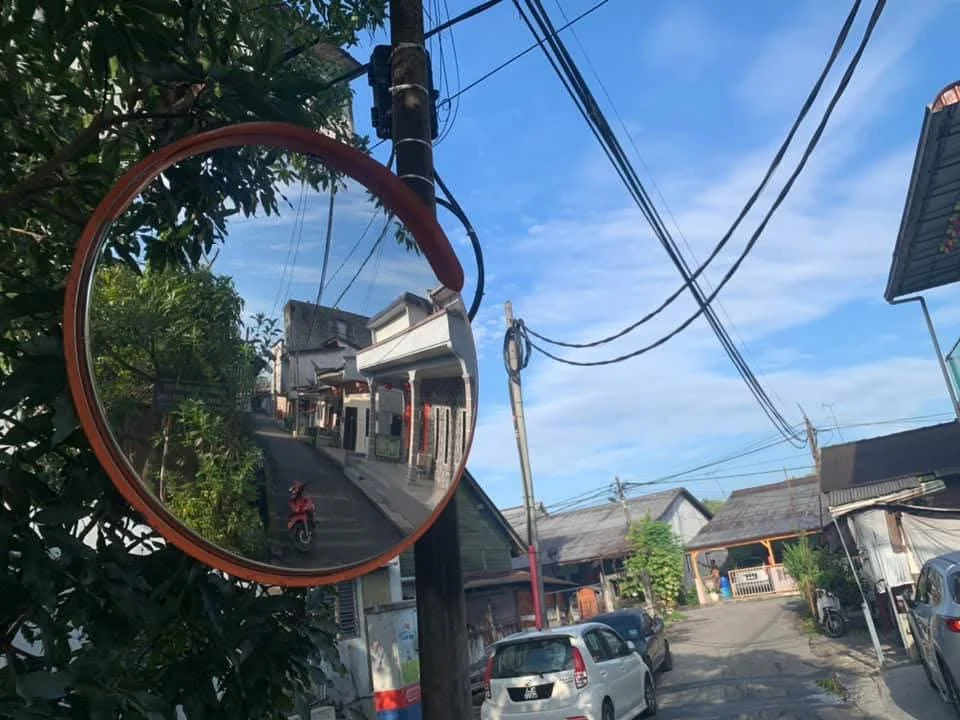 Specchio convesso con bordo arancione su un palo lungo una strada con abitazioni e veicoli.