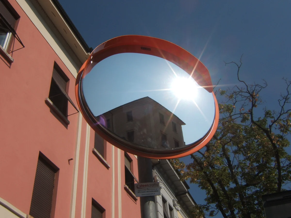 Specchio convesso con bordo arancione su un palo tra edifici e alberi, con il sole riflesso sulla superficie.